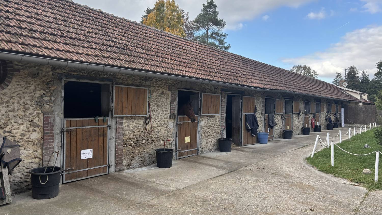 Façade en pierre et en bois des boxes, avec des chevaux à leur porte